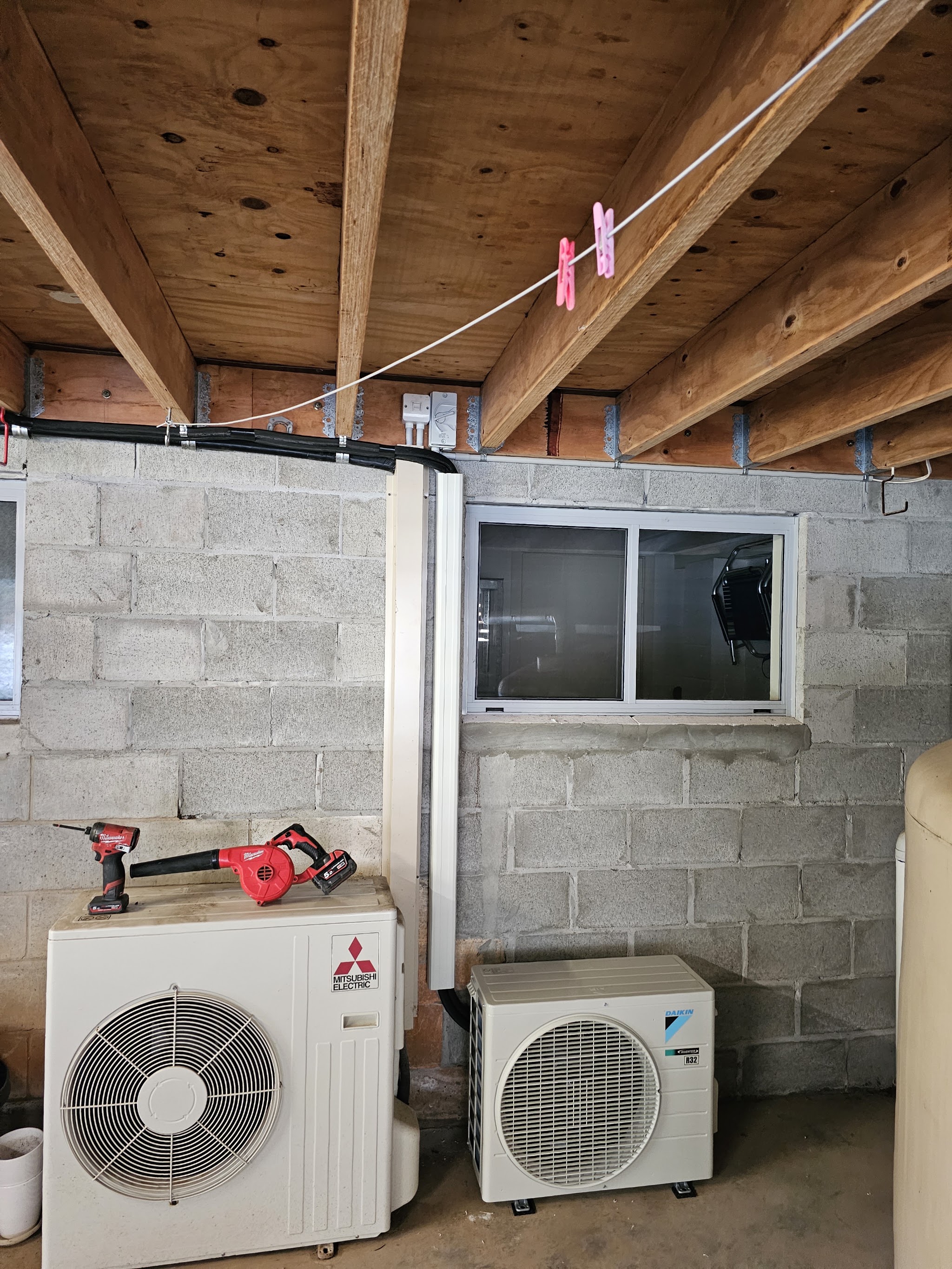 Two outdoor air conditioning units, a Mitsubishi Electric and a Daikin, installed under a high-set house with tools resting on top